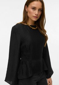 Melns pepluma tops ar garām piedurknēm, teksturētu audumu un savāktu vidukli. Salikts kopā ar apaļu krelliņu kaklarotu.