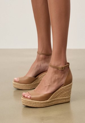 Picioare purtând sandale cu platformă în culoarea bronzului, cu curele la gleznă și talpă din funie împletită, pe un podea netedă bej.