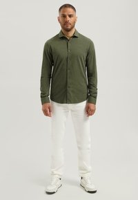 Olijfgroen overhemd met lange mouwen en knopen, van een geweven stof, gecombineerd met witte broek en witte sneakers. Minimalistisch ontwerp, met één logo op de borst.