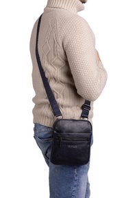 Borsa a tracolla in pelle nera con tracolla regolabile, forma rettangolare, tasca frontale con zip e dettaglio del logo in rilievo. Superficie strutturata.