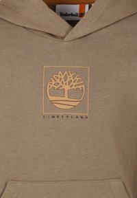 Felpa marrone con cappuccio, caratterizzata da un logo centrale di un albero beige e la parola "TIMBERLAND" in viola sotto. Tessuto morbido.