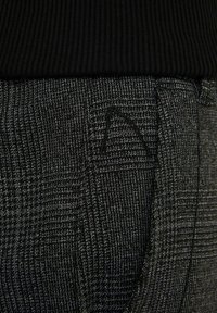 Detailní záběr na šedou a černou kostkovanou látku s kapsou a černým žebrovaným pasem, který zdůrazňuje texturu a detaily šití.