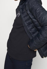 Mand iført navy blæserjakke over en sort sweatshirt med hvid skrift, hænderne i lommerne, og mørkeblå jeans mod en ensfarvet baggrund.