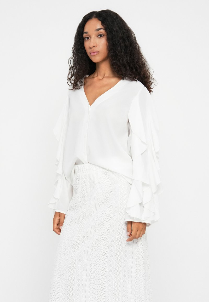 Weißes Blusenshirt mit V-Ausschnitt, Knopfverschluss und rüschigen Ärmeln. Kombiniert mit einem weißen strukturierten Rock, der komplizierte Muster aufweist.