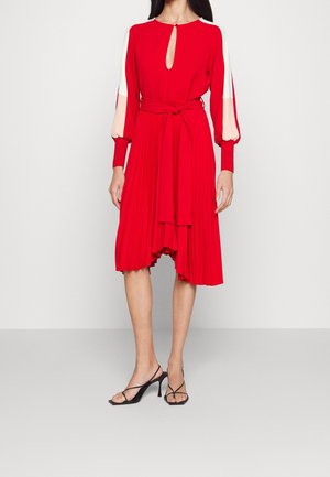 Robe plissée rouge avec un décolleté en trou de serrure, manches longues avec des accents blancs et une taille nouée ; assortie à des talons noirs à lanières.