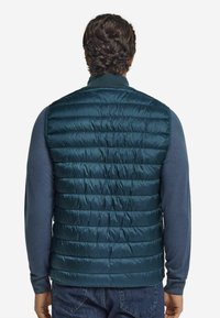 Tjock mörkt teal-väst med horisontellt quiltat mönster, ribbad krage och blå långärmad skjorta under, sedd bakifrån.