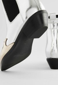 Botines plateados hasta el tobillo con elástico negro en los laterales, puntera afilada y tacón bajo negro. Presentan un acabado metálico suave y detalles en crema contrastantes.