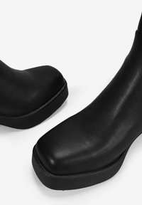 Botas de tobillo negras hechas de cuero suave con punta cuadrada y suela gruesa, presentando un diseño minimalista y sin herrajes visibles.