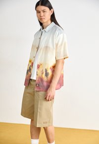 Short-sleeve shirt in cream with a horse graphic pattern in orange and purple; paired with beige shorts and white ankle socks.