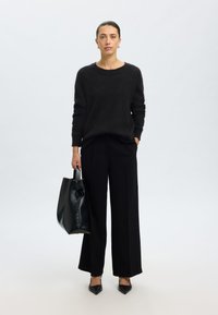 Camisola preta de malha com decote largo, combinada com calças pretas de perna larga feitas por medida. Os acessórios incluem uma mala de couro preta e sapatos de salto pontiagudos.