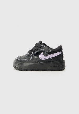 Sort Nike-sneaker med tyk sål, lilla Swoosh-logo, sorte snørebånd og hvide syninger, vist fra siden på en ensfarvet baggrund.