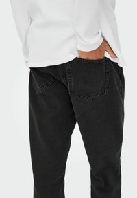 Pantalones vaqueros negros con un corte relajado, que cuentan con dos bolsillos traseros y una camisa de manga larga blanca texturizada metida en la cintura.
