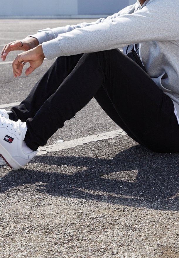 Zapatillas blancas con acentos rojos, pantalones negros ajustados y una sudadera gris, sentado sobre una superficie asfáltica texturizada.