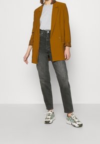 Hnědý dvouřadý blazer přes šedé tričko, doplněný černými vysokými džínami a vícebarevnými teniskami se zelenými akcenty.