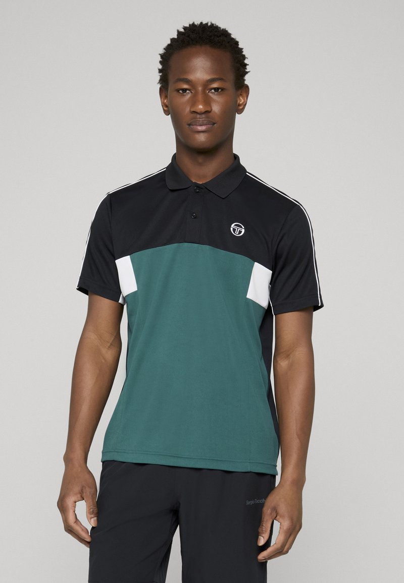 Fiatal férfi áll fekete és zöld poloingben, fehér csíkokkal az ujjakon és logóval a bal mellrészen, fekete nadrággal párosítva.