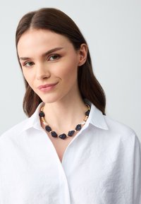 Collana di perle nere con pietre di forma irregolare e accentuazioni dorate, indossata sopra una camicia bianca con colletto. Texture liscia, design elegante.