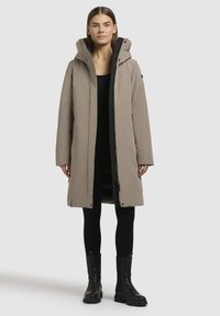 Lang beige gewatteerde jas met een capuchon, voorzien van een rits aan de voorkant en zijzakken, gedragen over een zwarte outfit en zwarte laarzen.