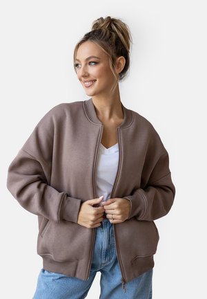 Brązowa rozpinana bluza z okrągłym dekoltem, w oversize'owym kroju, z prążkowanymi mankietami i bocznymi kieszeniami, noszona na białą koszulkę i niebieskie dżinsy.