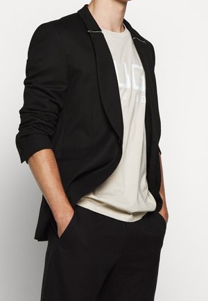 Homme portant un blazer noir aux manches retroussées sur un t-shirt graphique beige et un pantalon noir, les mains dans les poches, sur un fond uni.