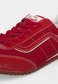 Червена маратонка Vans с бели шевове и лого, червени връзки и кафява гумена подметка, показана от страничен ъгъл.