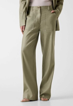 Mujer con pantalones anchos de color verde oliva, sandalias beige con tiras y una blazer verde oliva a juego sobre una camiseta de canalé rosa claro.