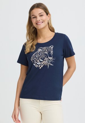 Mujer sonriente que lleva una camiseta azul marino con un gráfico de cara de leopardo, combinada con pantalones beige claro, posando contra un fondo liso.