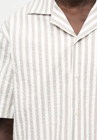 Άνδρας που φοράει πουκάμισο μπεζ και λευκό με κάθετες ρίγες, κοντά μανίκια, μυτερό γιακά και κουμπιά μπροστά.