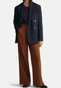 Frau in navy-blauem, doppelt geknöpftem Blazer, navy-blauem Kabelstrickpullover, weiten braunen Hosen und dunkelbraunen Schuhen, die mit einer Hand in der Tasche posiert.