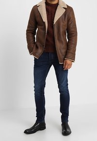 Brun fusk läderjacka med en krämfärgad fårskinnskrage, bärs över en vinröd stickad tröja och blå jeans, matchat med svarta ankelstövlar.