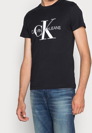 Mężczyzna w czarnym T-shircie Calvin Klein Jeans i niebieskich dżinsach stoi na jednolitym jasnym tle.