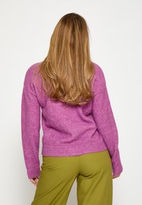 Violet vilnas džemperis ar ribotu apakšmalu un piedurknēm, kombinēts ar olīvu zaļām biksēm. Audums ir mīksts ar strukturētu virsmu.