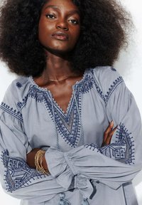 Blouse gris clair avec une broderie complexe en bleu marine, un ourlet en dentelle et des manches bouffantes dotées de poignets élastiques. Franges décoratives à l'ourlet.