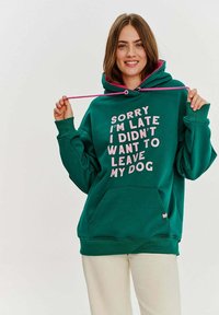 Zelená bavlněná mikina s růžovými stahovacími šňůrkami a velkým textem "Omlouvám se, že jsem pozdě, nechtěl jsem opustit svého psa." Součástí je přední kapsa.