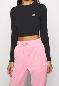 Černý zkrácený top s dlouhým rukávem a třemi šedými pruhy na rukávech, s bílým logem Adidas. V kombinaci s světle růžovými sametovými teplákovými kalhotami.