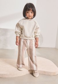 Sudadera color crema con cuello redondo, combinada con pantalones de pierna ancha a rayas en tonos beige y marrón claro. Completado con zapatillas de tono marrón claro sobre un fondo claro.