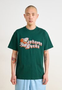 T-shirt en coton vert avec un canard cartoon et un graphique de basketball ainsi que le texte "Carhartt Ducks" en orange et blanc, coupe décontractée.