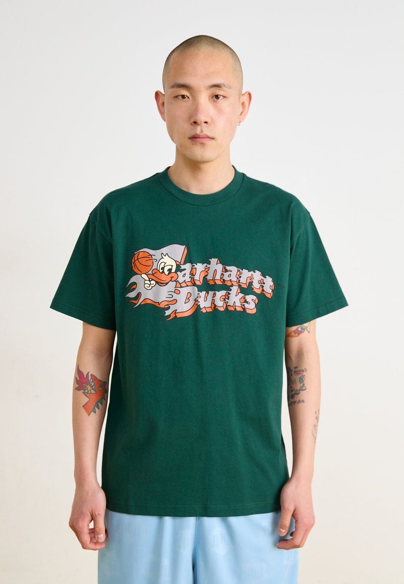 T-shirt en coton vert avec un canard cartoon et un graphique de basketball ainsi que le texte "Carhartt Ducks" en orange et blanc, coupe décontractée.
