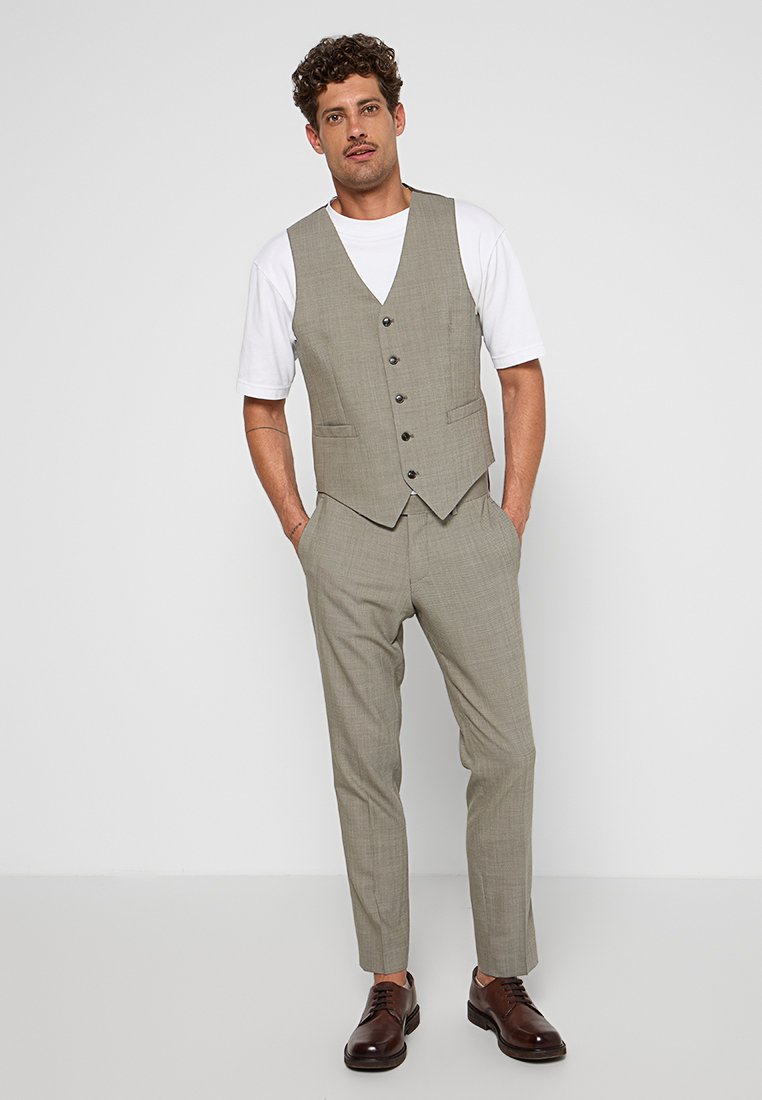 Gilet gris clair avec cinq boutons, associé à un pantalon tailleur assorti. T-shirt blanc en dessous, complété par des chaussures en cuir marron.