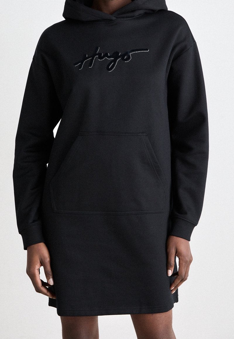Robe sweat à capuche noire avec poche kangourou et logo en cursive sur la poitrine, portée par une personne à la peau foncée sur un fond uni.