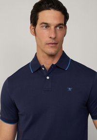 Marinblå polo-shirt med texturerad tyg, ljusblå krage-accenter och en liten broderad logga på vänster bröst.