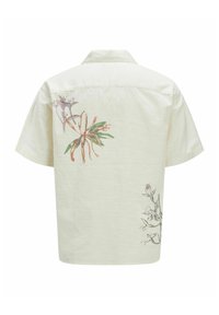 Camisa de manga corta de color beige claro con cuello. Presenta patrones florales en verde, rosa y púrpura en la parte de atrás con un acabado texturizado.