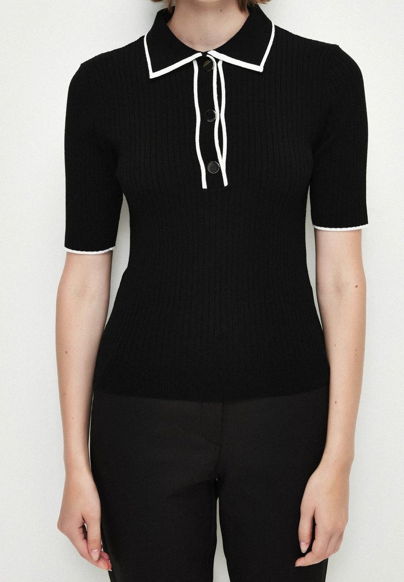 Polo noir en maille côtelée avec manches courtes, col blanc et détails de boutons, avec un bord contrastant blanc le long des bords.