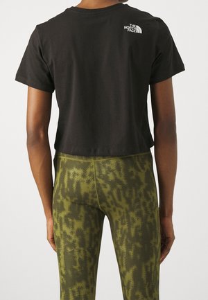 Person iført en sort cropped t-shirt med The North Face-logo og grønne mønstrede leggings, set bagfra mod en ensfarvet baggrund.