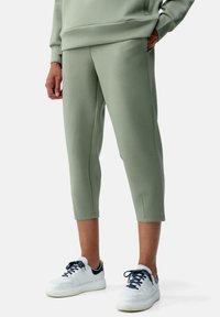 Pantaloni sportivi corti di colore verde chiaro realizzati in stoffa liscia, dotati di tasche laterali, abbinati a sneaker bianche con dettagli e lacci blu marino.