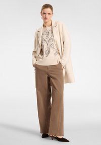Blazer beige sobre sudadera beige con patrón negro, combinado con pantalones anchos marrones y zapatos de tacón marrones. Tela suave, corte relajado.