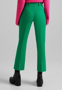 Femme portant un pantalon taille haute vert, des bottes à plateforme noires, et un pull en côte rose vif, se tenant contre un fond gris uni.