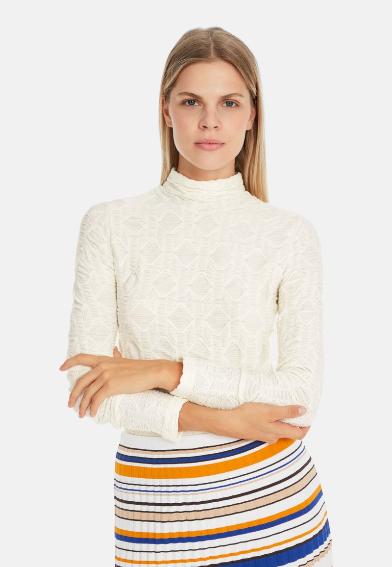 White patterned long-sleeve top with a high neck, featuring textured geometric designs and ruffled cuffs, paired with a striped pleated skirt.