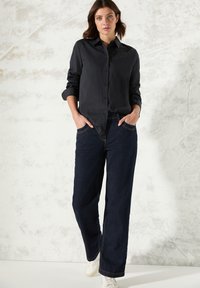 Camicia nera con bottoni e colletto, con dettagli di borchie su spalle. Abbinata a jeans a gamba larga blu scuro e sneakers bianche.