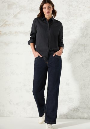 Camicia nera con bottoni e colletto, con dettagli di borchie su spalle. Abbinata a jeans a gamba larga blu scuro e sneakers bianche.