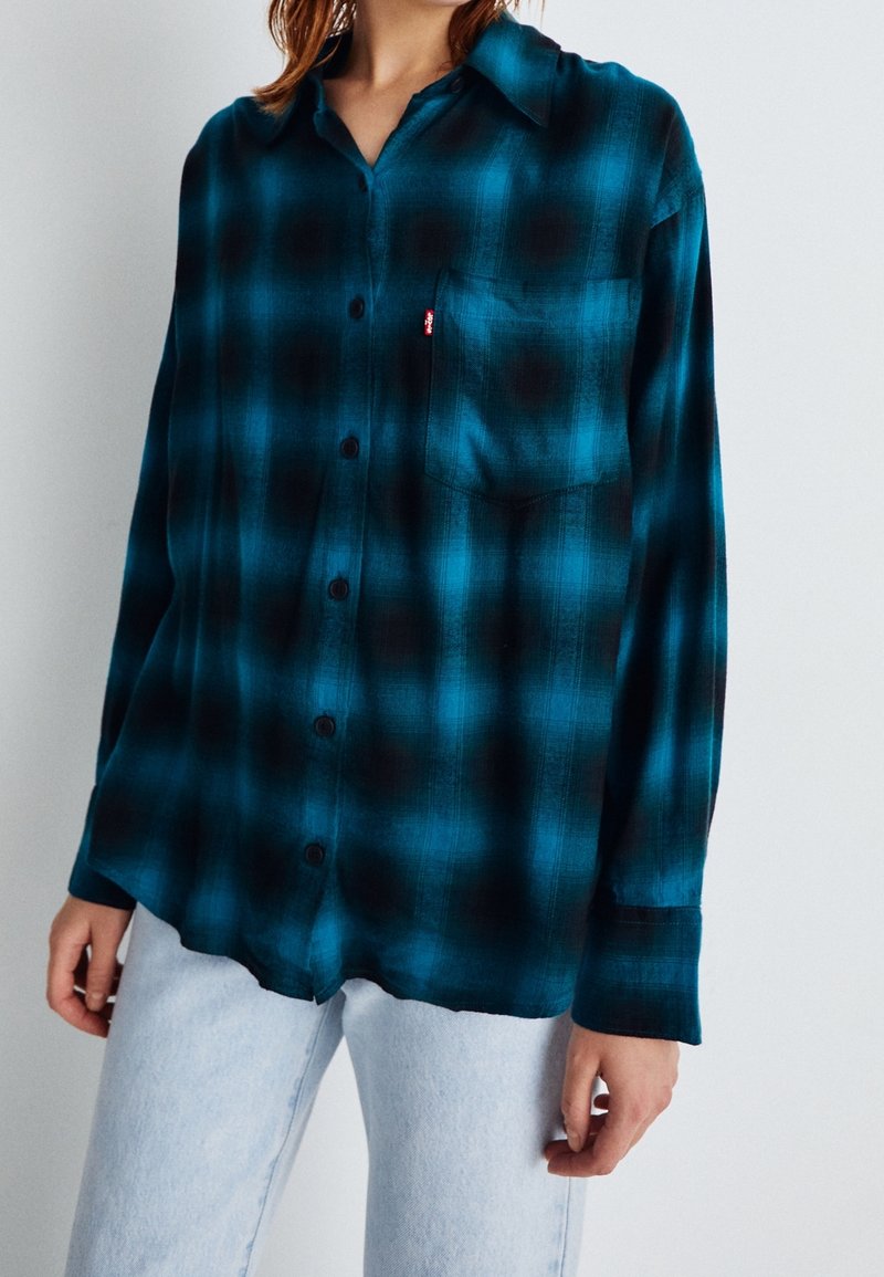 Femme portant une chemise ample à carreaux bleu sarcelle et noir avec poche poitrine, et un jean bleu clair.